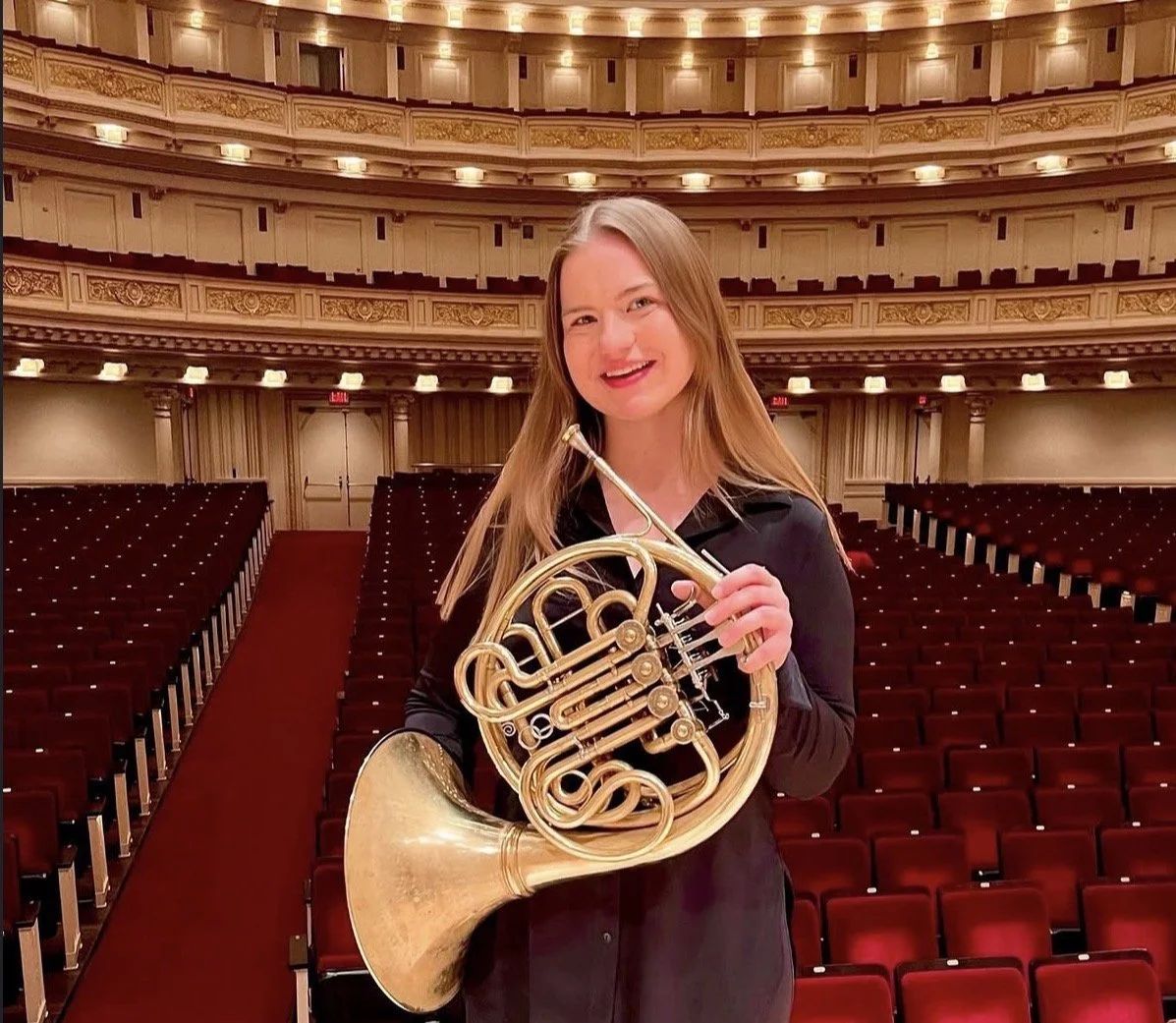 Young woman standing in an empty orchestra hall holding a French horn.