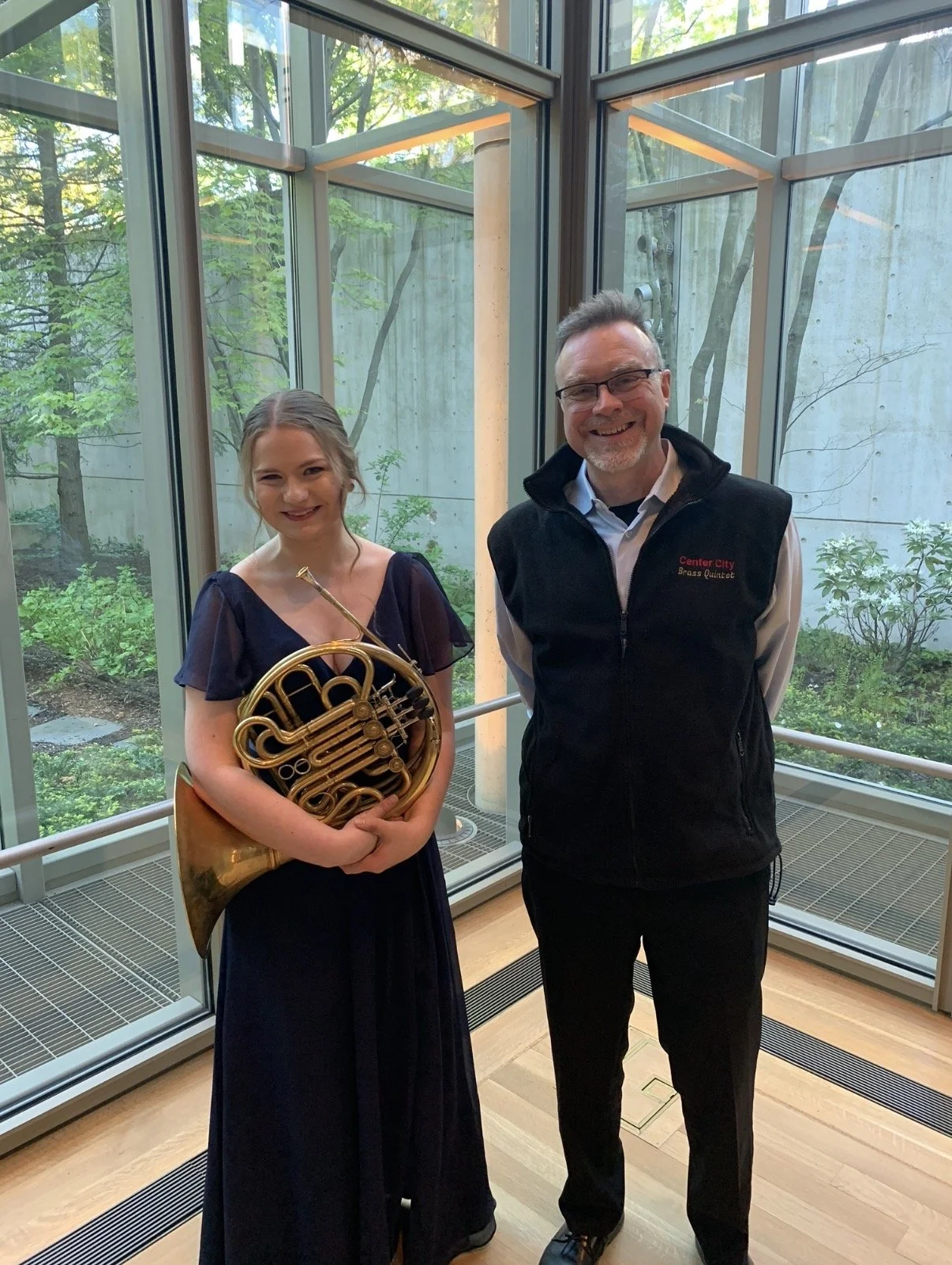 A young woman in a dark blue dress holding a French horn, standing next to a man in a black vest with glasses, both smiling inside a modern building with large glass windows and greenery outside.