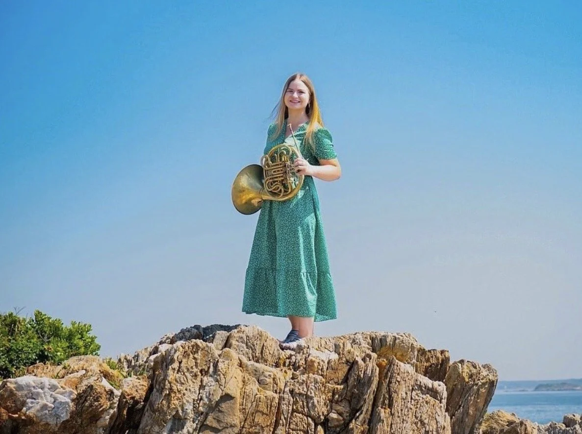 A young woman in a green dress standing on rocky terrain at the coast, holding a French horn, smiling under a clear blue sky.