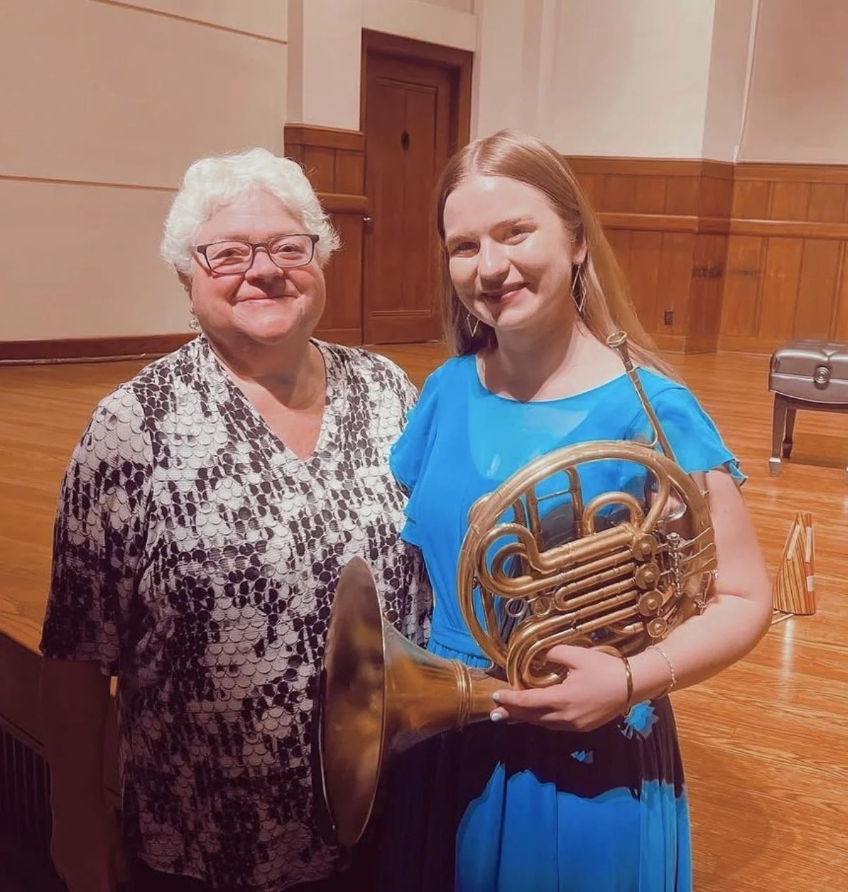 Two women smiling, standing on a wooden stage with a wooden door in the background. One woman has short white hair, glasses, and is wearing a patterned blouse. The other woman has long red hair, is holding a French horn, and is wearing a bright blue dress.