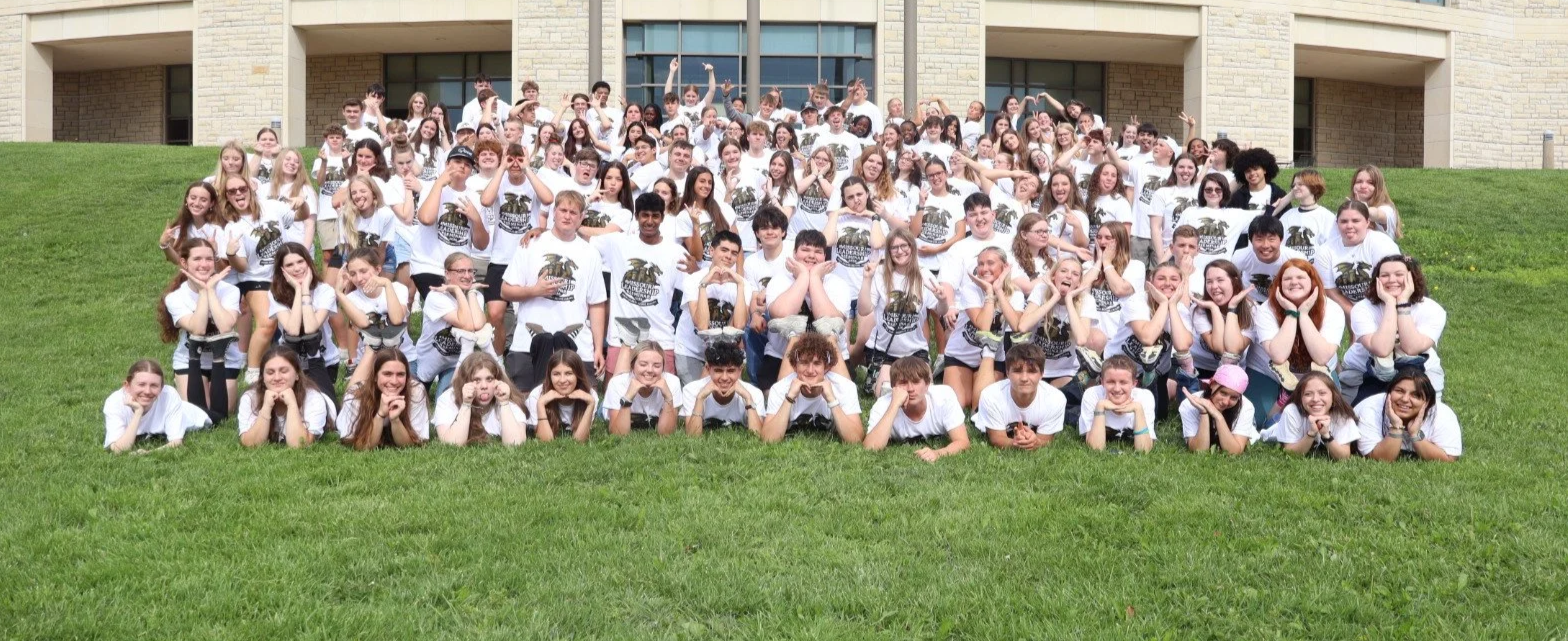 Large group of students posing on a grassy lawn outside a school building, wearing matching white T-shirts.