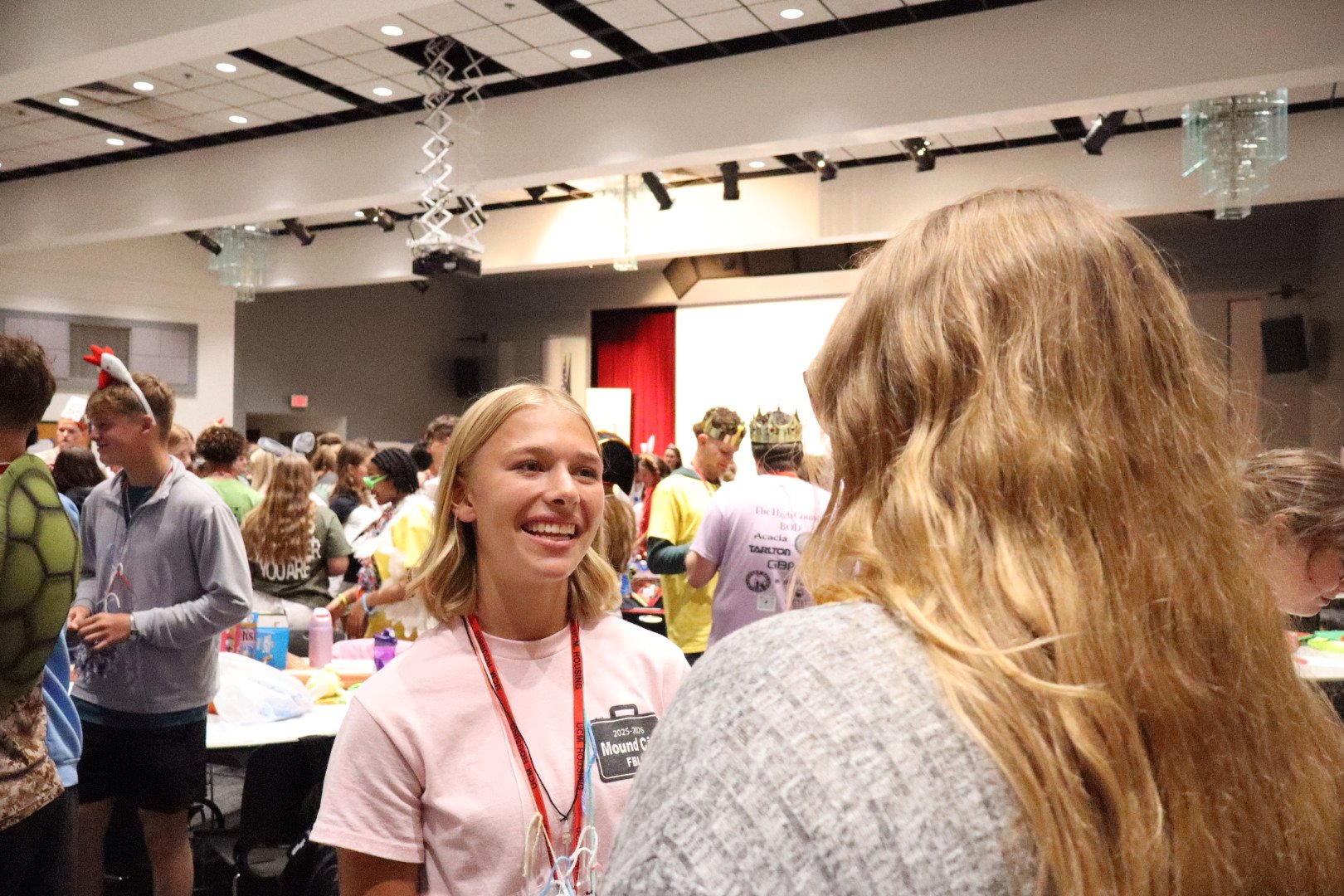 Smiling young woman wearing a pink shirt and event lanyard at a crowded indoor gathering, with attendees and a stage in the background.