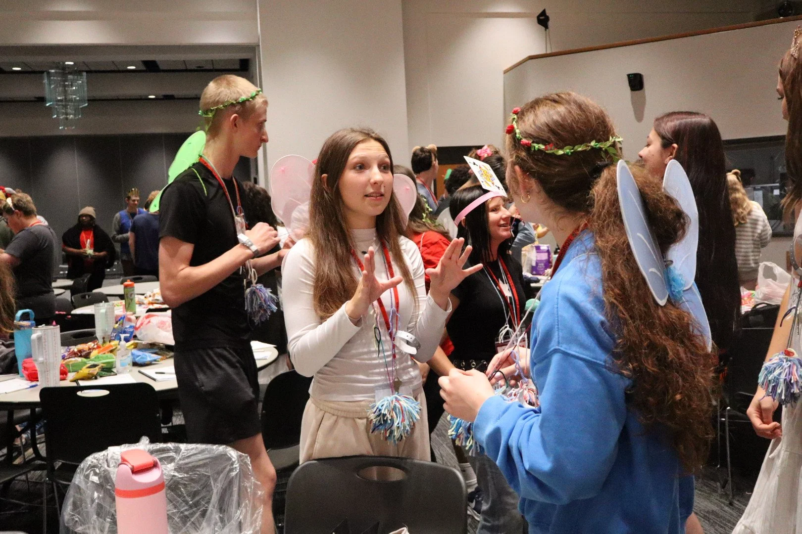 People in colorful fairy costumes and wings socialize at an indoor event.