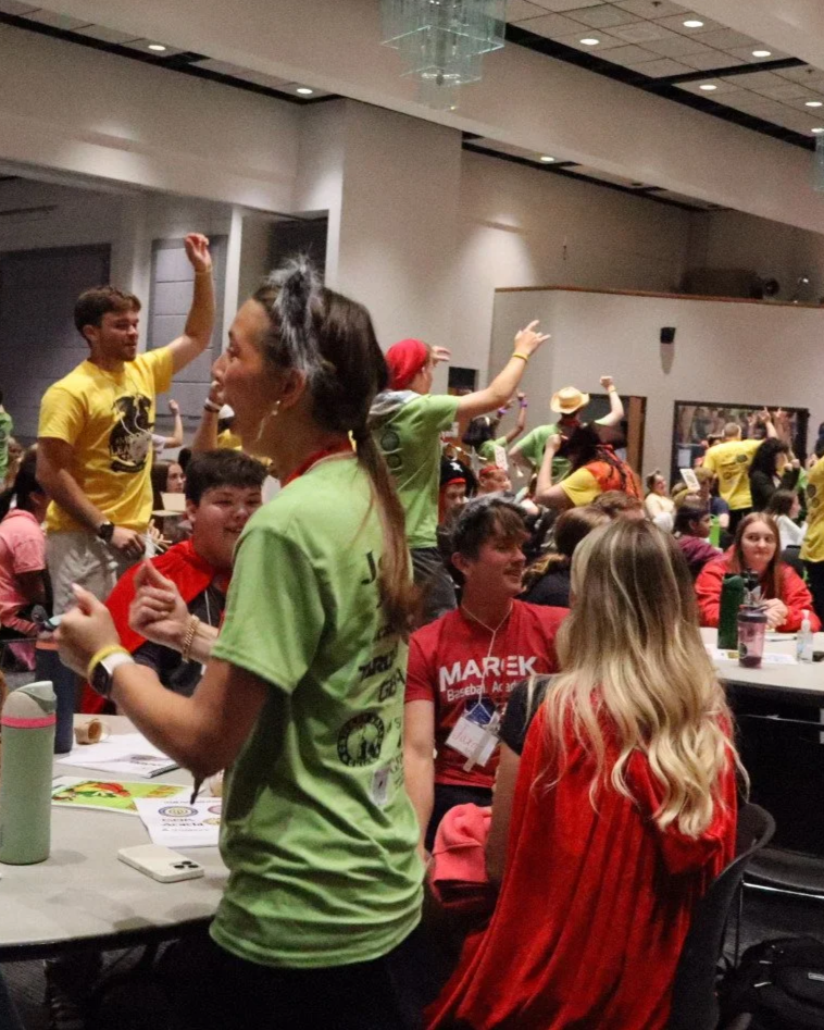 Crowded conference room with people in colorful event T-shirts standing, dancing, and socializing around tables.