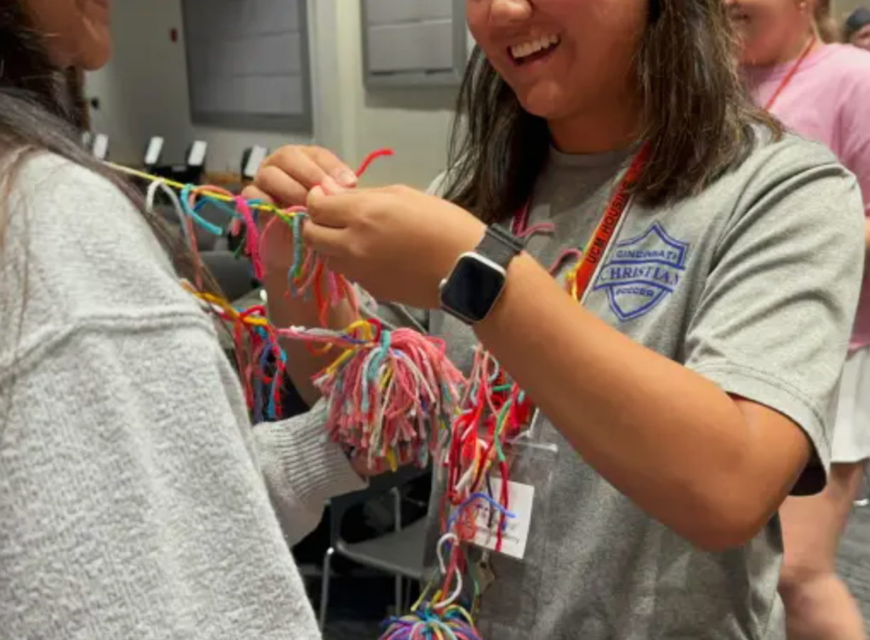 Smiling woman in a Girl Scouts T-shirt crafts colorful friendship bracelets with another person.