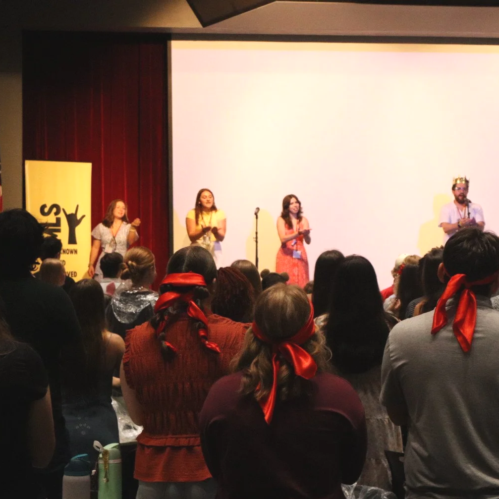 Audience watches four presenters onstage in front of a projection screen at an event, with several people wearing red ribbons.