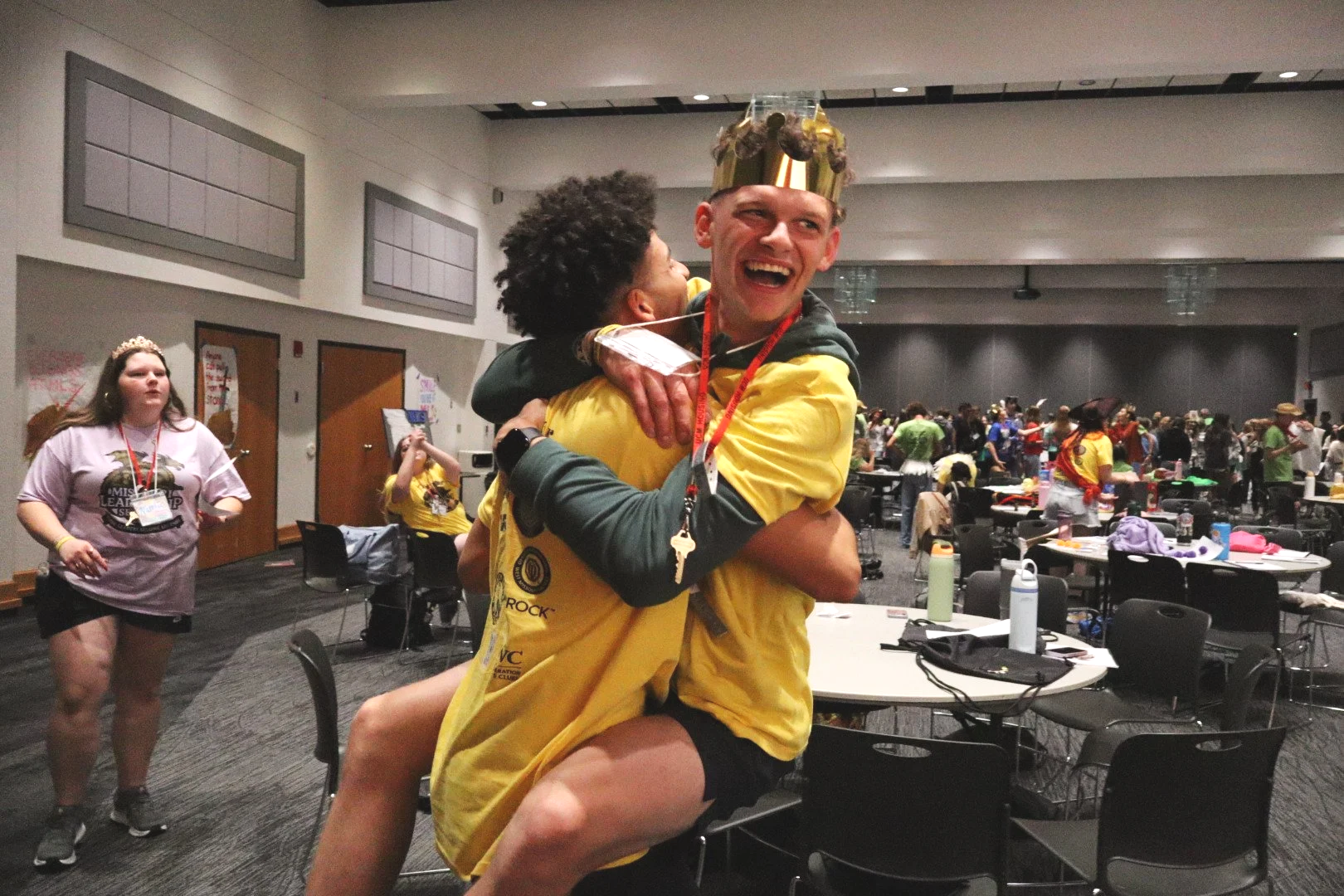 Smiling event participants hug in a crowded conference room as one person wears a shiny gold crown.