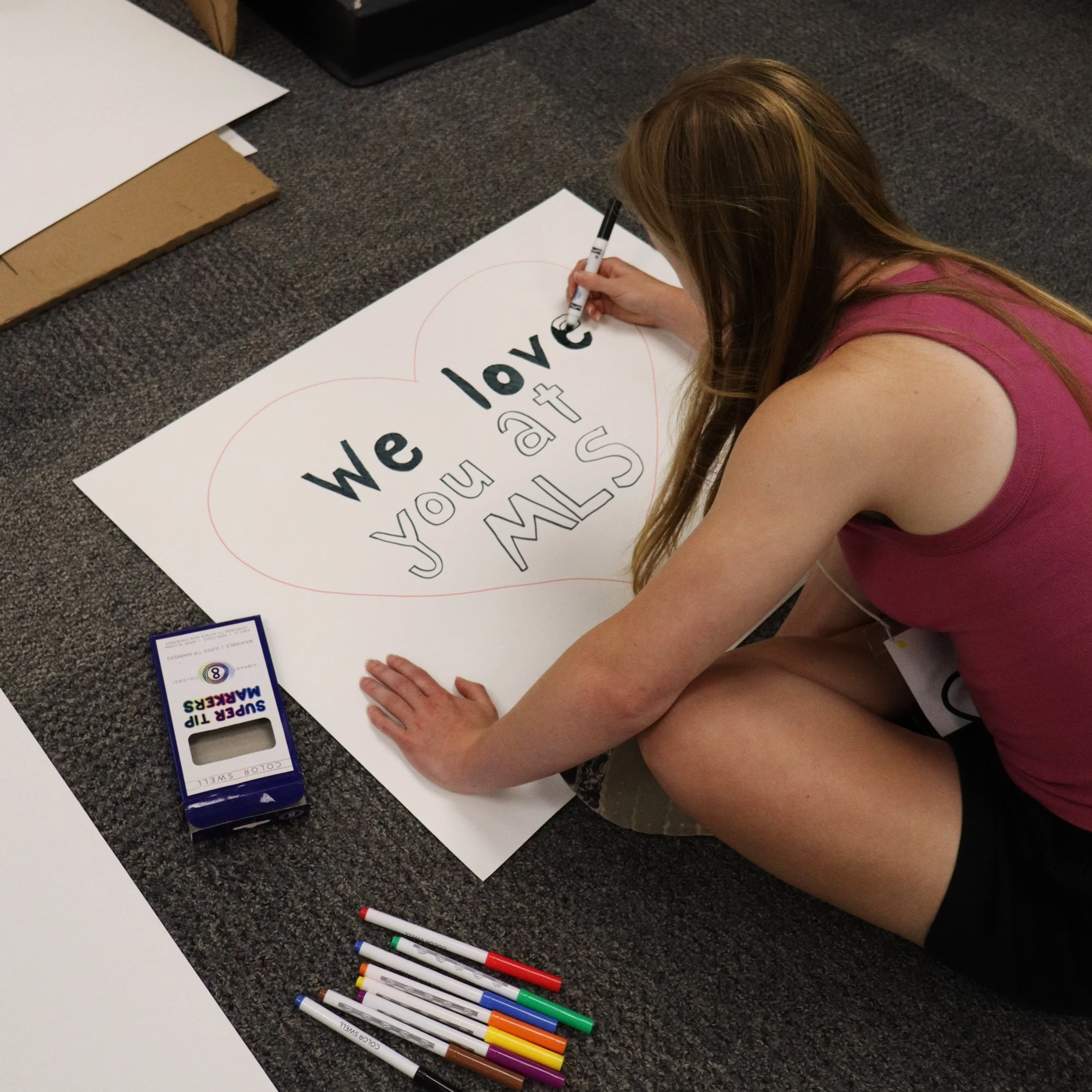 Woman drawing a large heart and lettering on poster board with colorful markers.