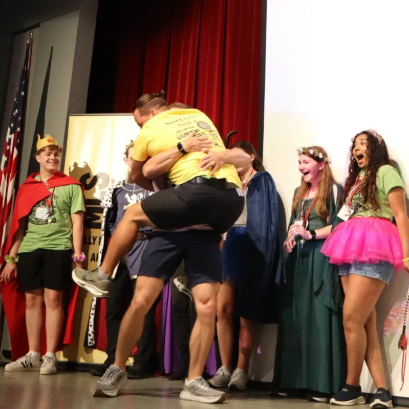 Costumed performers celebrating onstage beside red theater curtains, with one person lifting another in a hug.