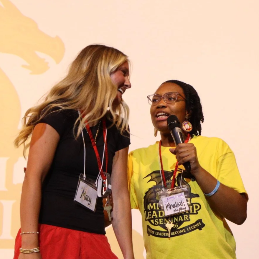 Two women wearing event badges stand onstage; one holds a microphone while the other leans in to speak.