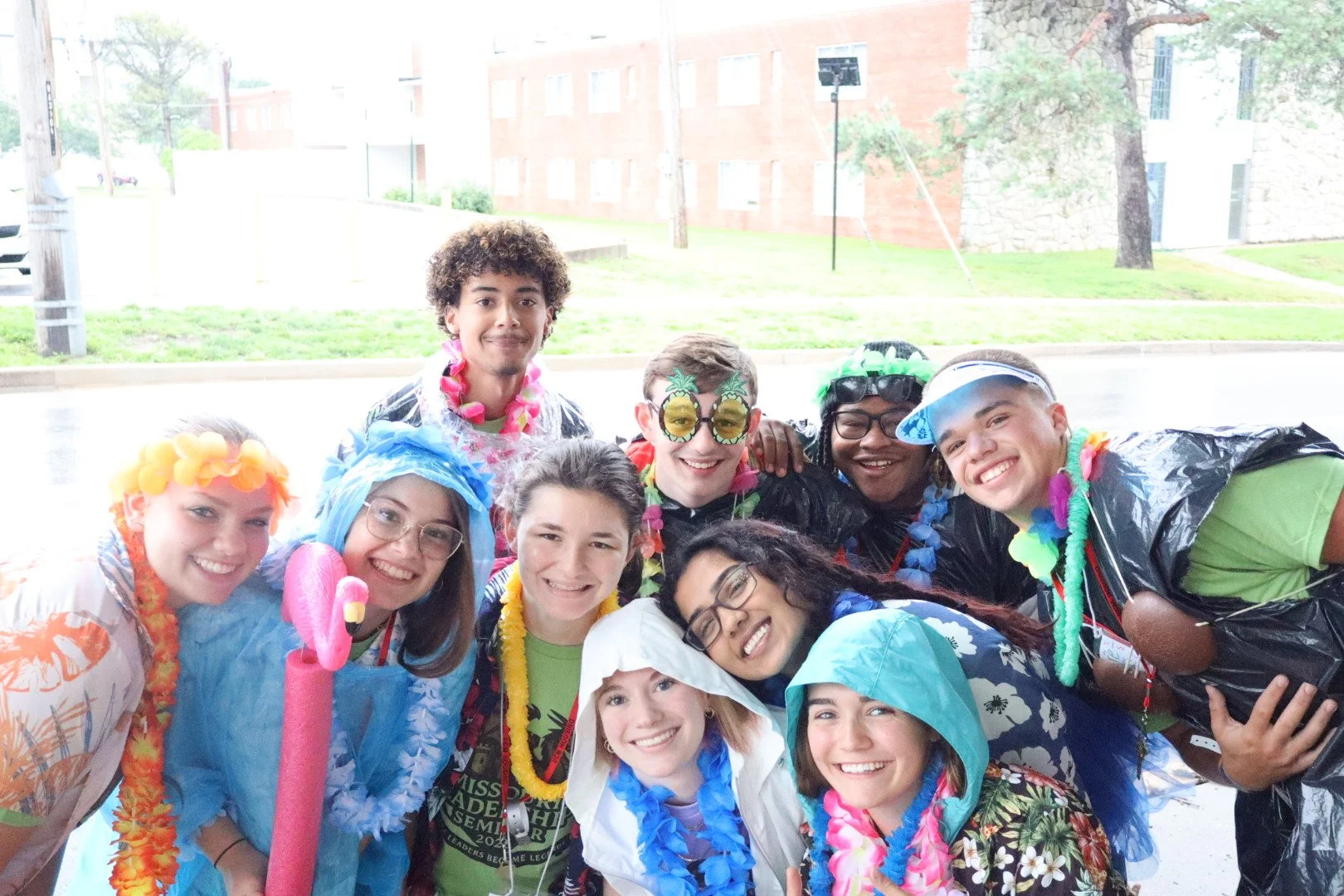 Group selfie of young people wearing colorful leis, costumes, sunglasses, and rain ponchos outdoors.