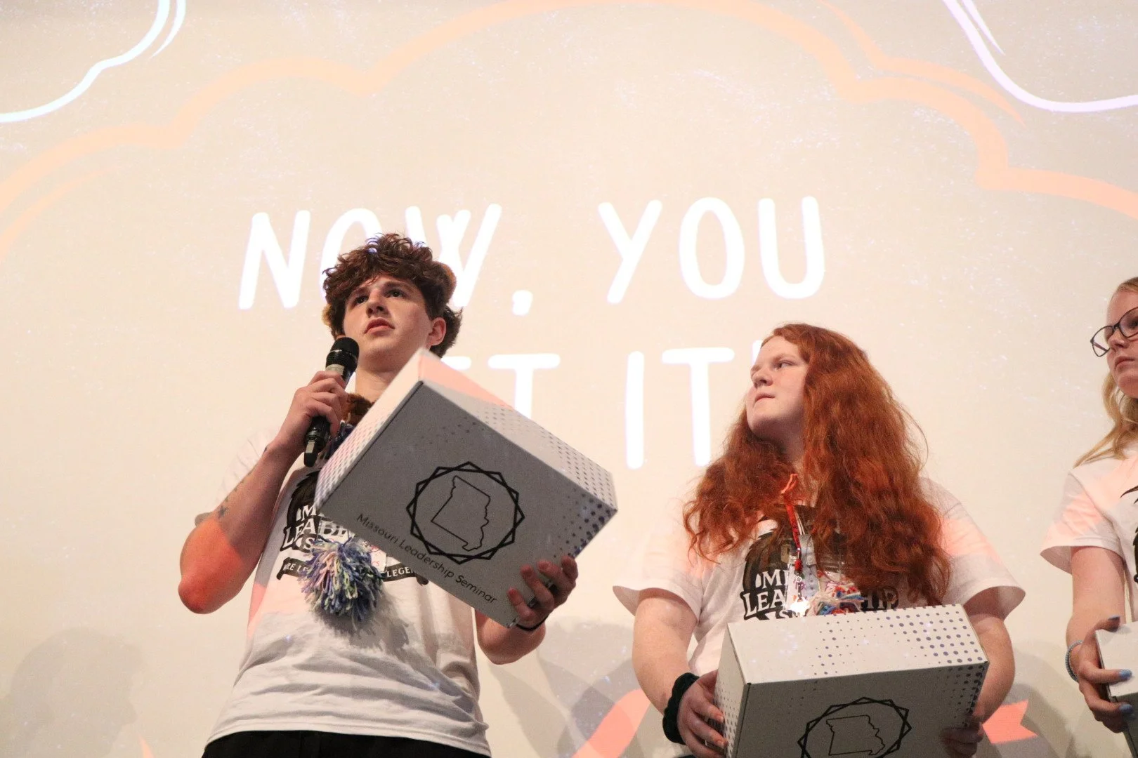 Youth leadership seminar participants onstage holding award boxes, with one speaking into a microphone in front of a projected presentation.