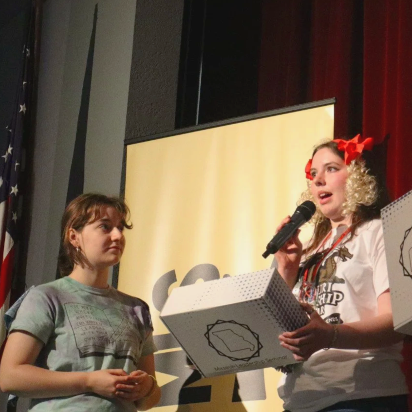 Woman speaking into a microphone beside another woman on a theater stage with an American flag and presentation screen.