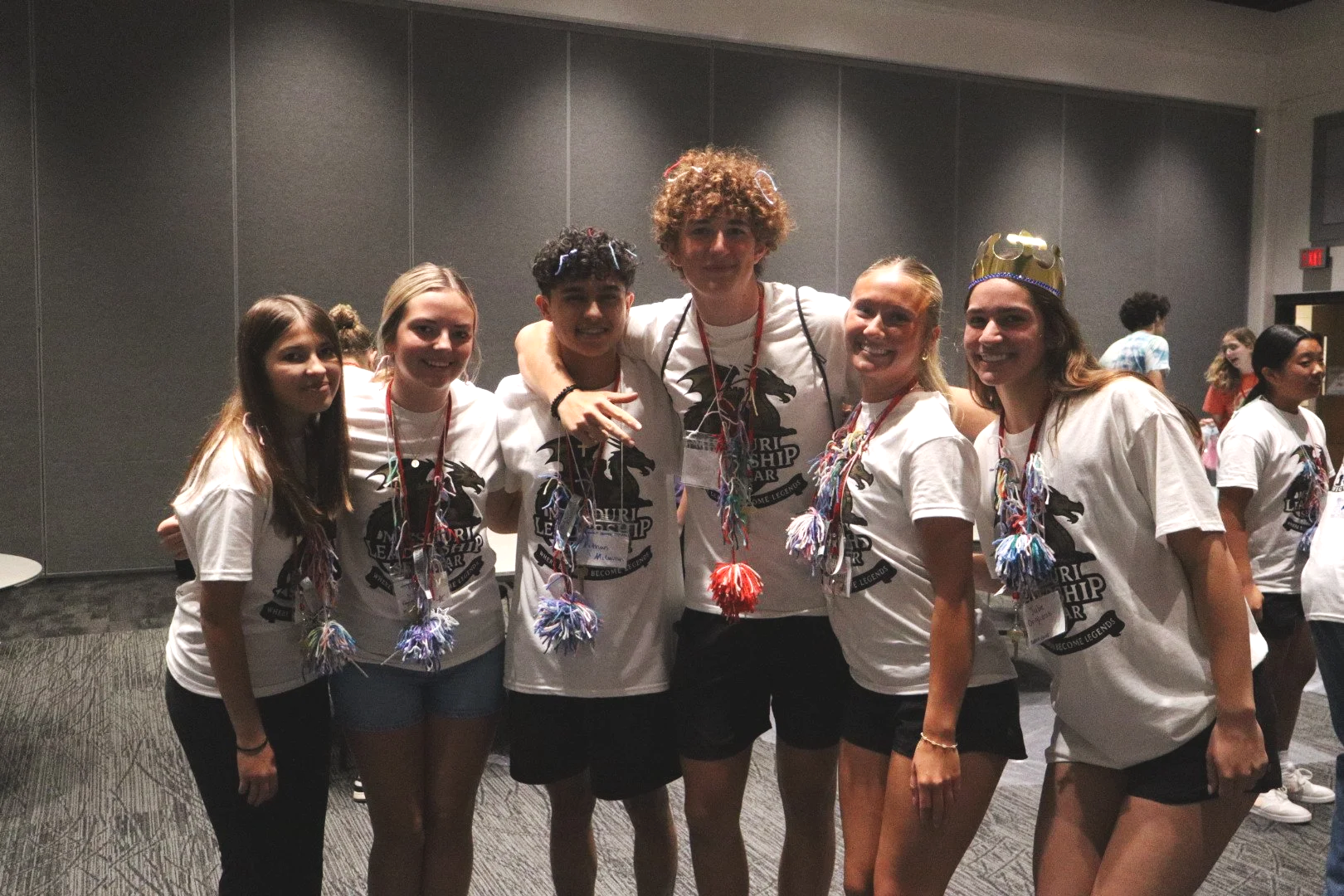 Group of young people wearing matching white shirts, colorful leis, and a crown at an indoor event