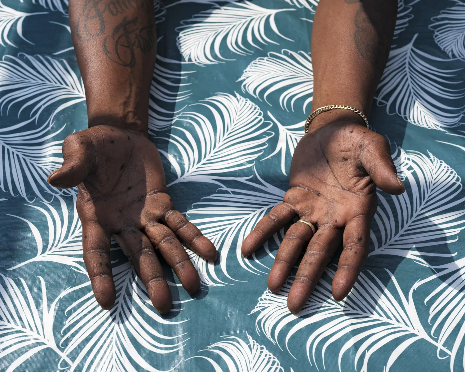 Close-up of outstretched hands with dirt and ink stains, resting on a blue surface with white palm leaf pattern.