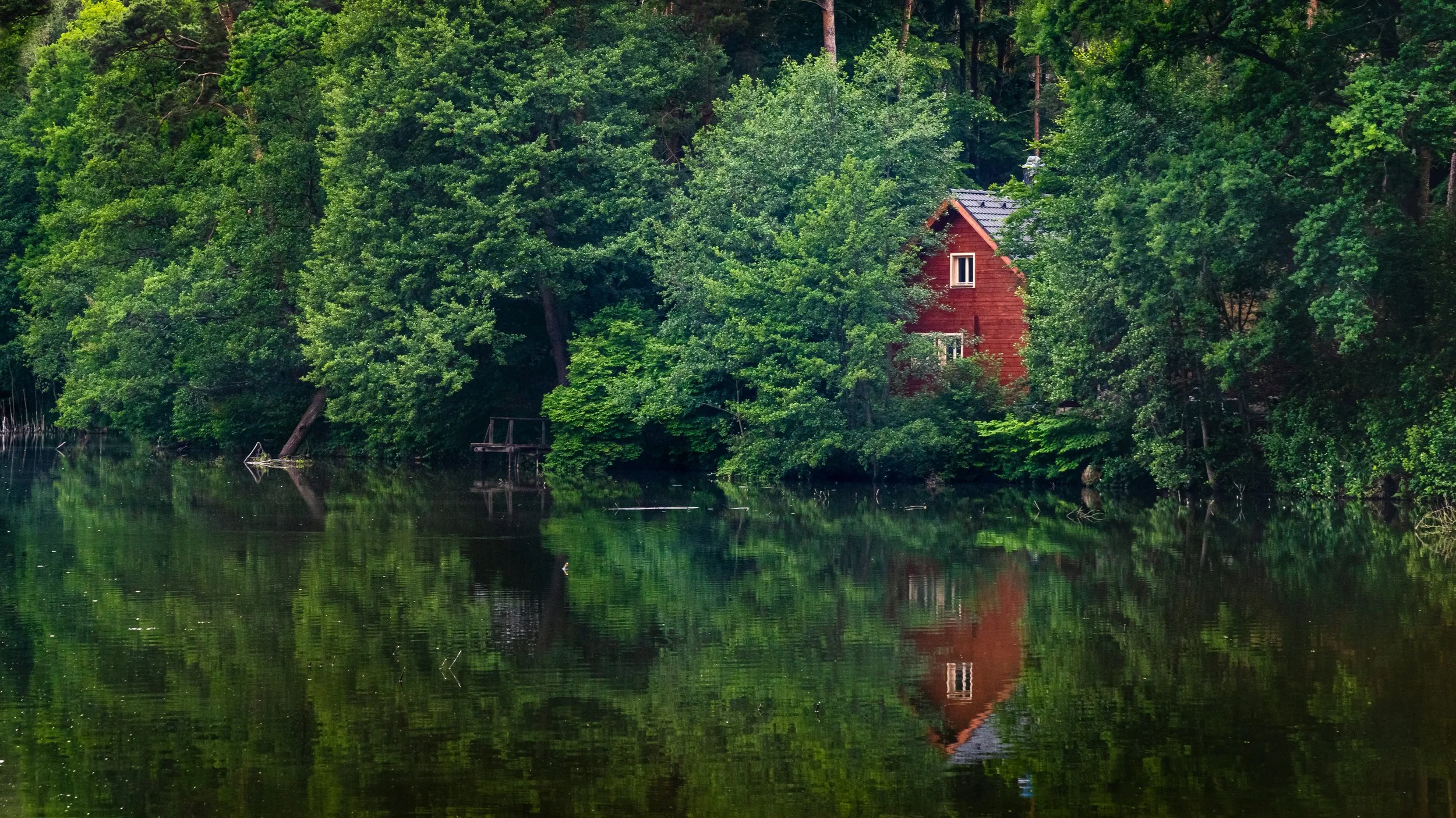 A red house surrounded by dense green trees beside a calm lake that reflects the scene.