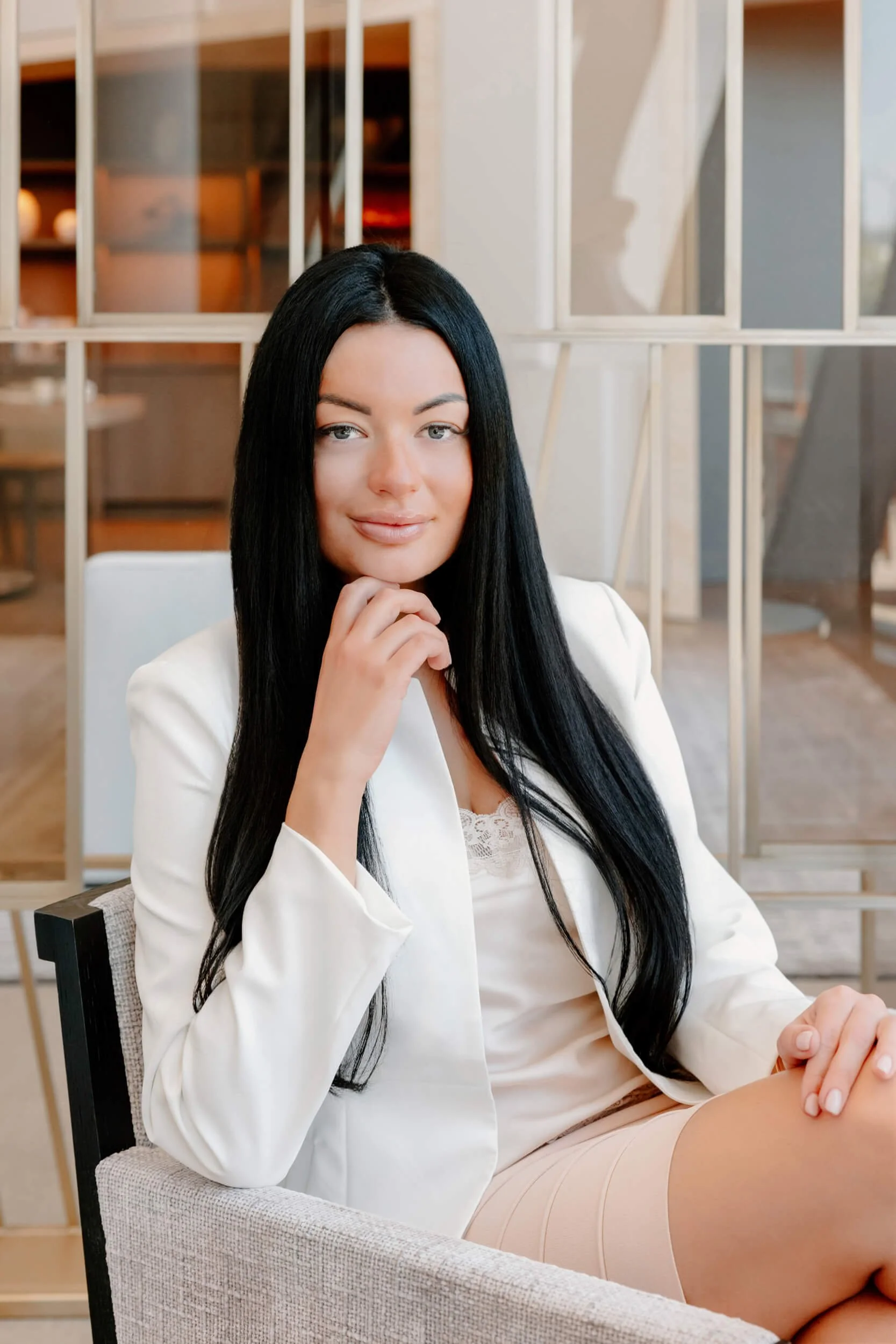 A woman with long black hair, wearing a white blazer and beige skirt, sitting in a modern, well-lit space with wood and glass decor, touching her chin with her hand and smiling at the camera.