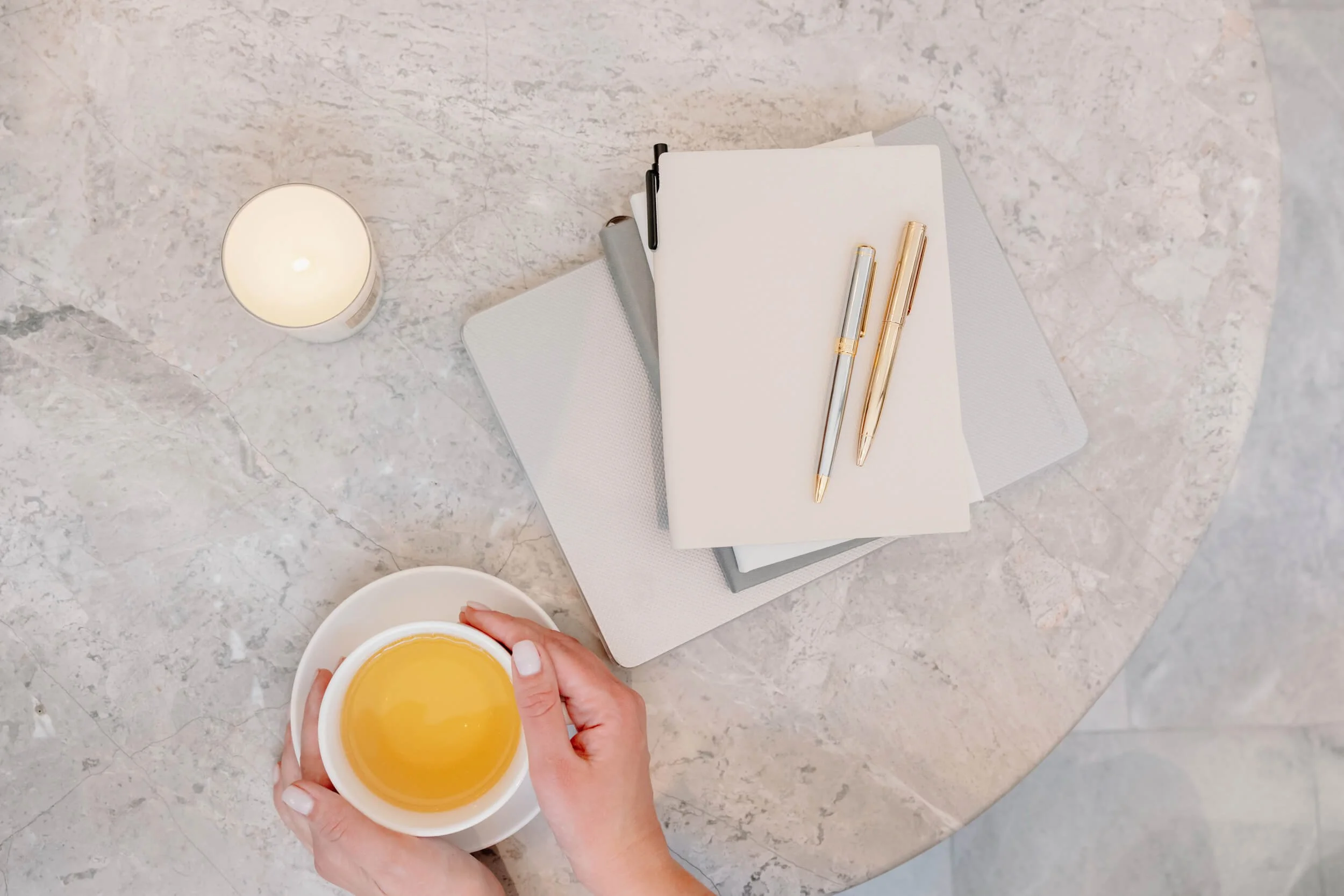 A person holding a cup of yellow liquid, with a workspace on a grey marble table that includes notebooks, pens, a cup of coffee, and a candle.