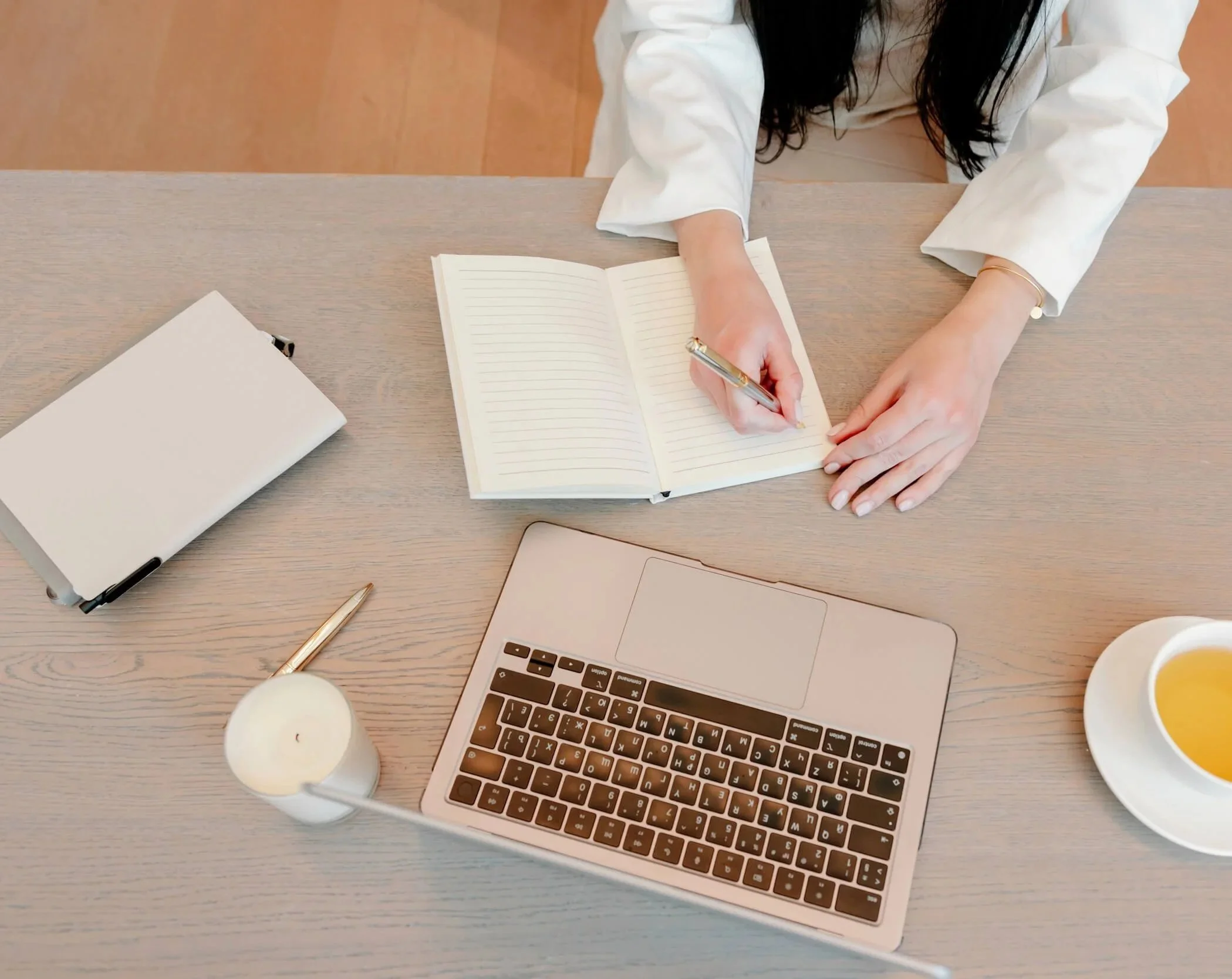 Top-down view of a person writing in a lined notebook at a wooden desk, surrounded by a laptop, candles, a cup of tea, and a closed laptop.