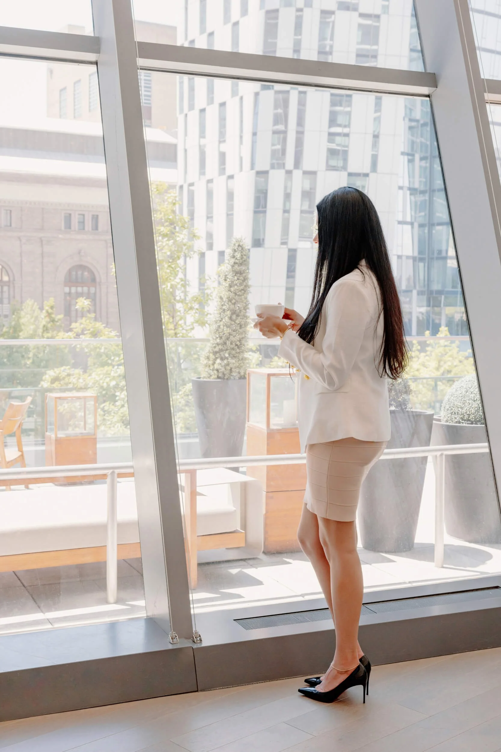 A woman in a white blazer and beige skirt is standing indoors near large glass windows, holding a white cup. She is wearing black high heels and has long black hair. The background shows a cityscape with modern buildings and some greenery.