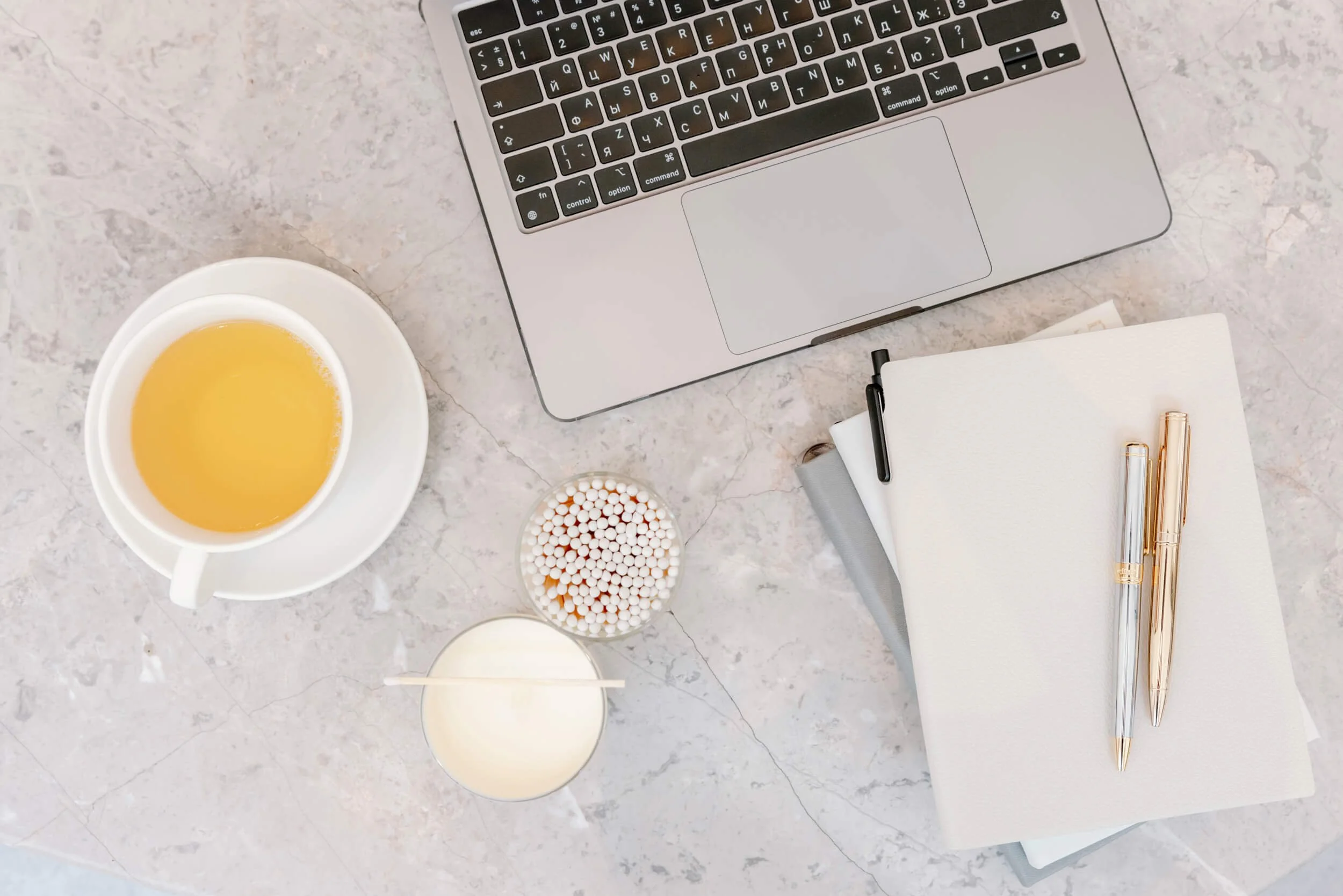 A top-down view of a workspace with a silver laptop, a cup of yellow tea on a white saucer, a small dish of white and orange candies, a white notebook, a black pen, and two gold pens on a light marbled surface.