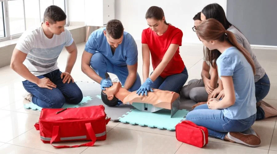 Group of people practicing CPR on a mannequin during a training session, with medical equipment nearby and a trainer demonstrating.