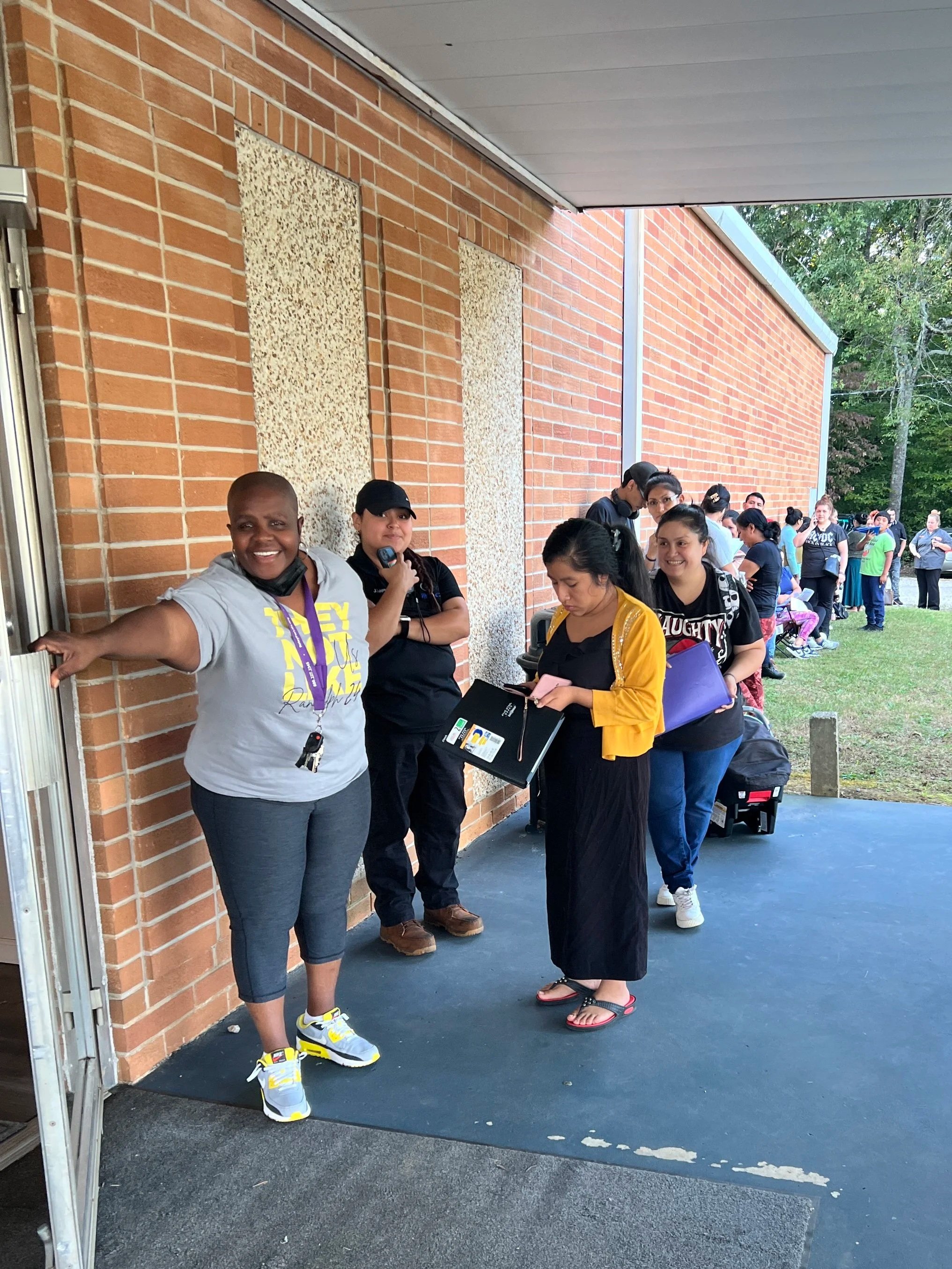 A line of people standing outside a brick building, waiting in line. The people are engaged in activities like talking, looking at their phones, or smiling. The weather appears to be clear and pleasant.