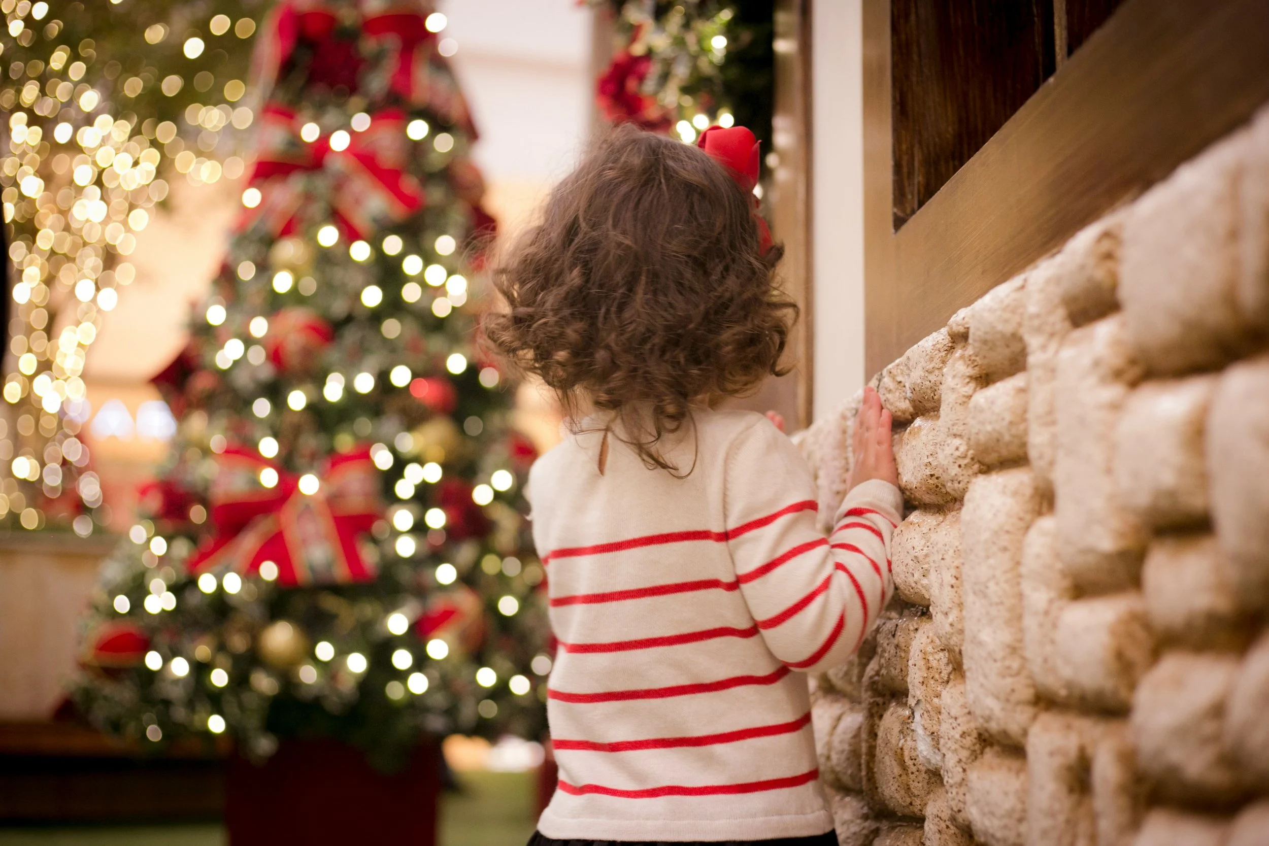 A young girl with curly hair wearing a beige sweater with red stripes, touching a stone fireplace, with a decorated Christmas tree with lights and red ribbons in the background.