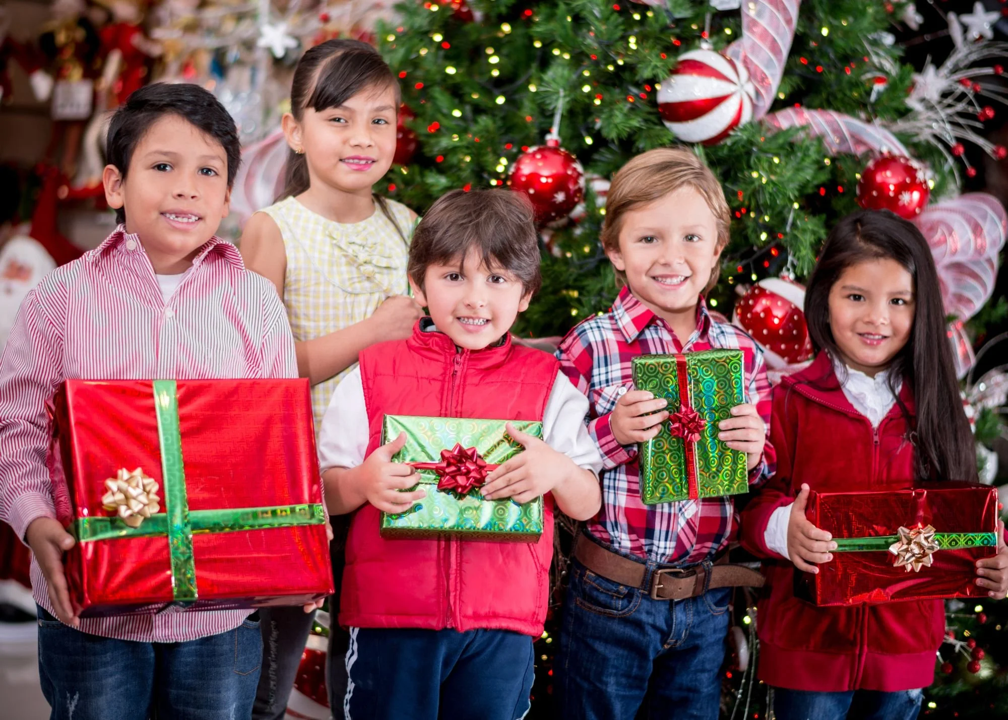 Group of five children holding Christmas presents in front of a decorated Christmas tree.