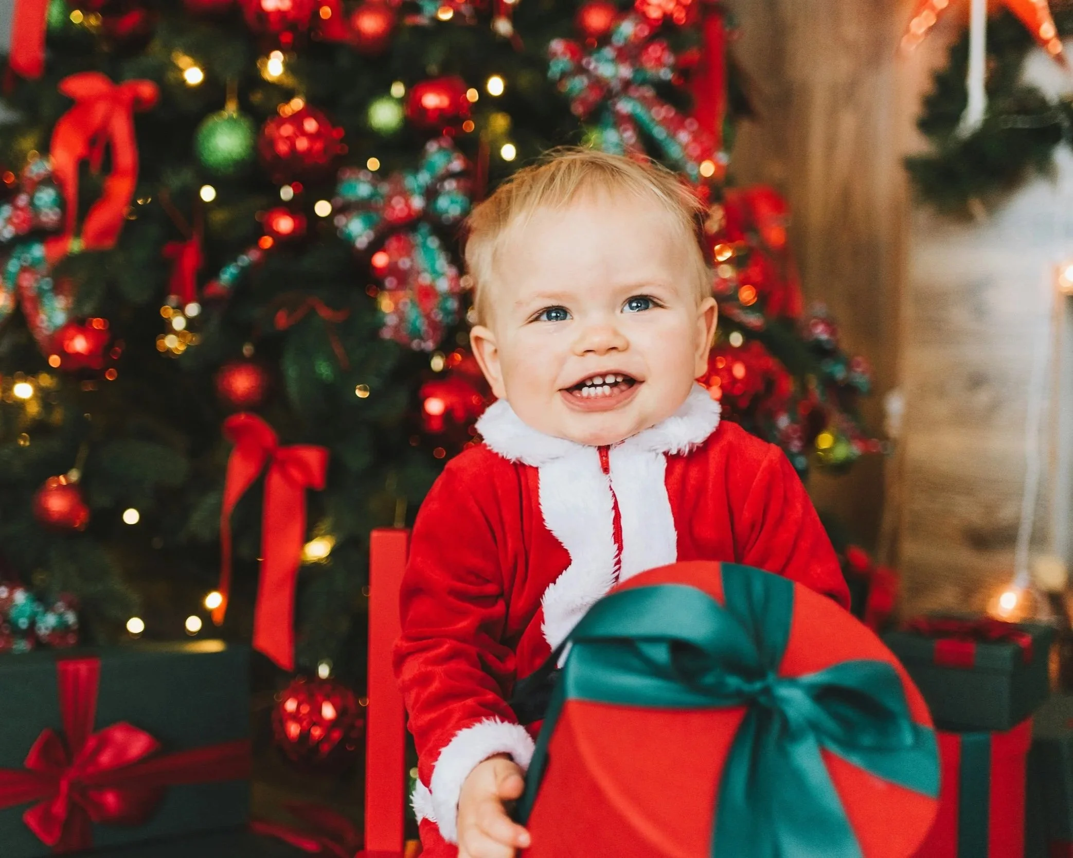 A young child with blonde hair and blue eyes, smiling, dressed in a red and white Santa outfit, sitting in front of a decorated Christmas tree with red and green ornaments and blurred warm lights.