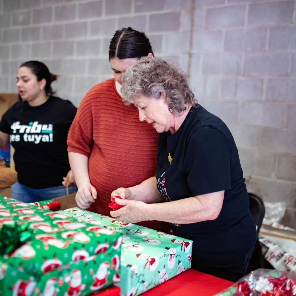 An elderly woman and a younger woman are wrapping Christmas presents at a table covered with holiday-themed wrapping paper and gifts.