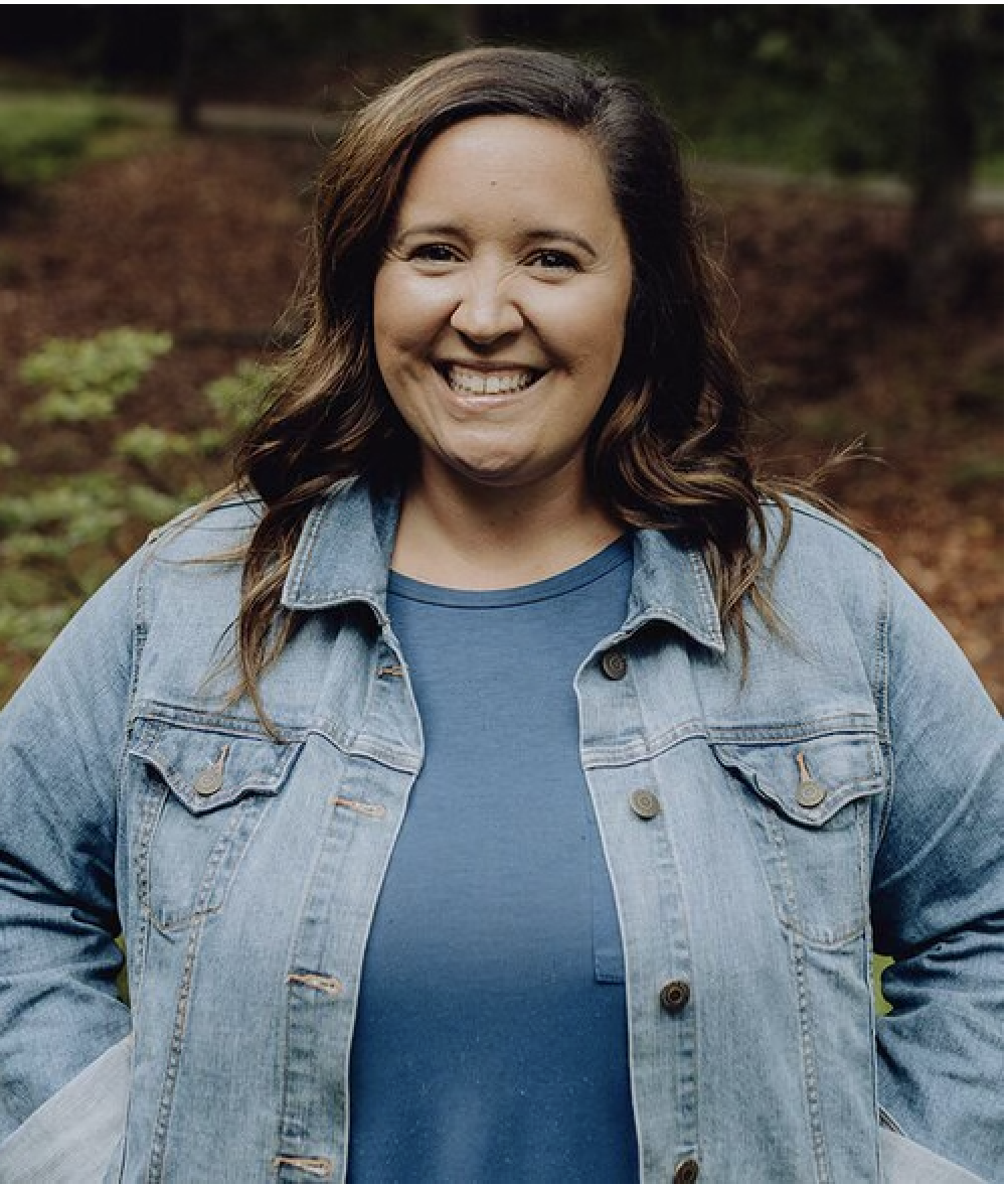A woman with shoulder-length dark hair, smiling, wearing a blue shirt and a light denim jacket, standing outdoors in a wooded area.