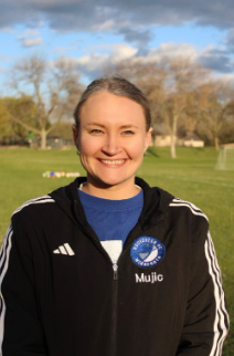 Young smiling woman outdoors, wearing a black sports jacket with a circular patch on the chest and a blue shirt underneath, in a park with trees and grass.