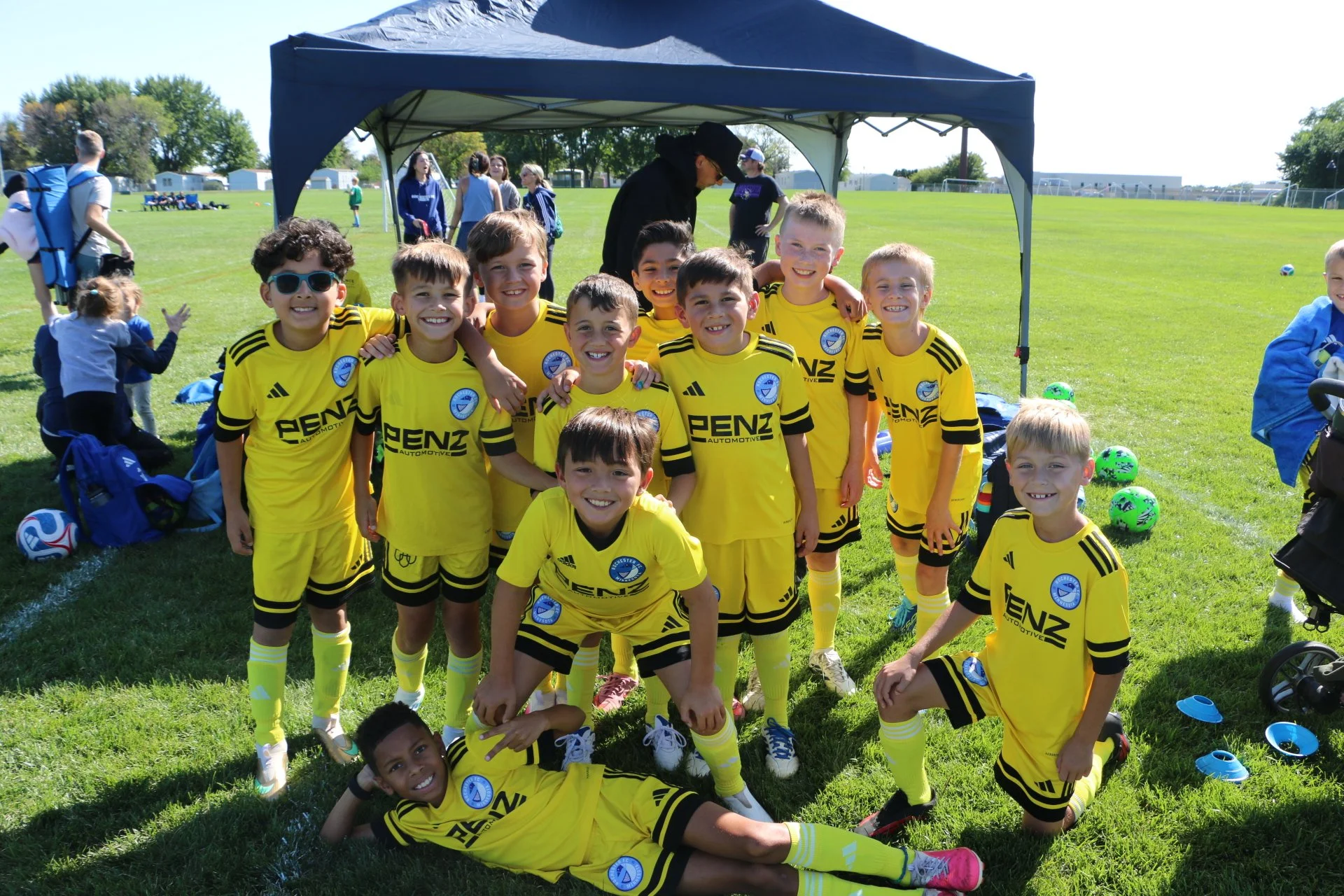 Group of young boys in yellow soccer uniforms posing outdoors on a sunny day, with some kneeling and others standing, under a canopy, on a grassy field with soccer balls and equipment nearby.