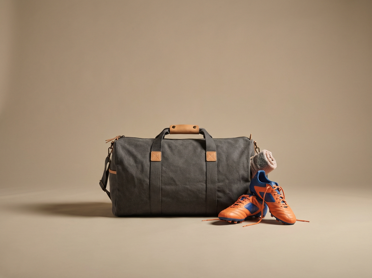 A gray sports duffel bag with tan accents, orange and blue soccer cleats, and a gray sock, on a beige background.