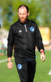 Man in black sportswear with a blue and white emblem on the chest, walking outdoors on a grassy field with trees in the background.