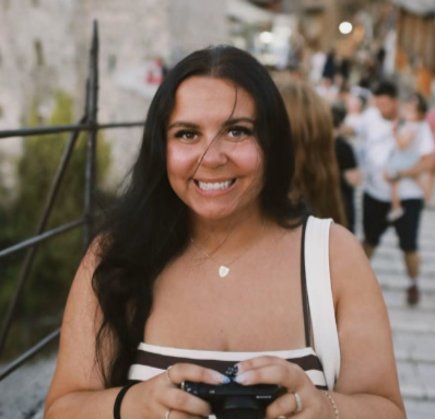 Smiling woman with long dark hair holding a phone, standing outdoors on a wooden bridge with people in the background.