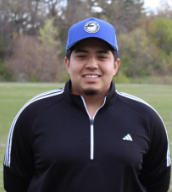 Young man in a blue baseball cap and black jacket standing outdoors on a grassy field.