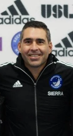 A man smiling, wearing a black jacket with the Adidas logo and the word 'SIERRA' on it, standing in front of a backdrop with Adidas and USL logos.