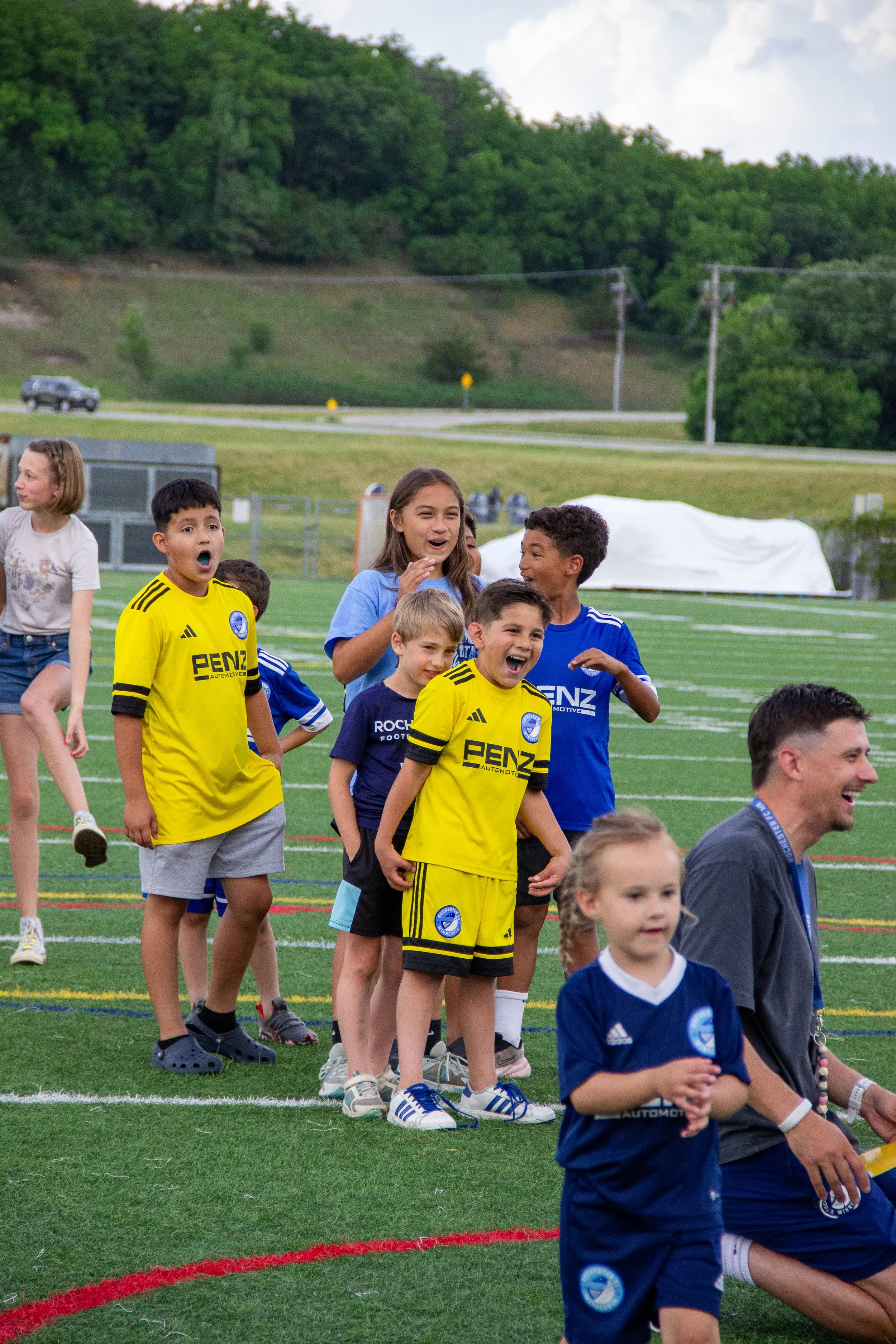Kids and adults on a soccer field, some in team jerseys, smiling and standing in line.