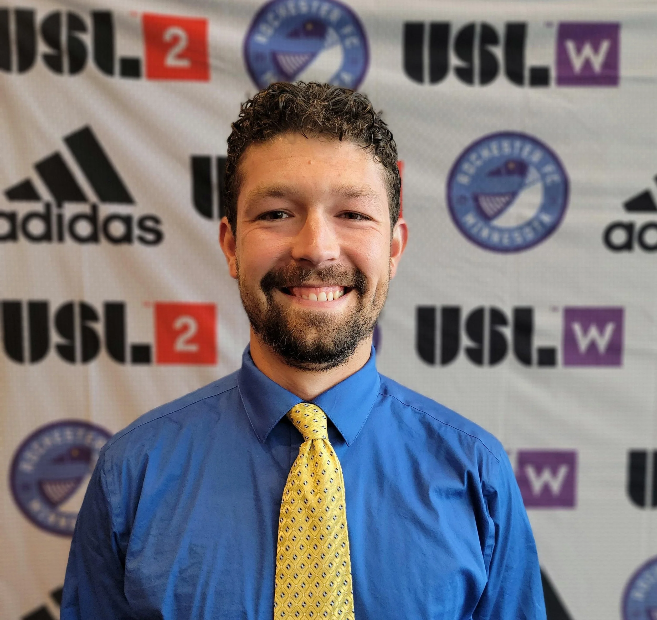 Young man with curly dark hair and a beard smiling, wearing a blue shirt and a yellow patterned tie, standing in front of a backdrop with various logos and the words 'U.S. Youth'.