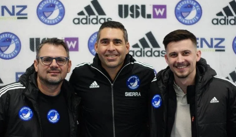 Three men standing together, wearing black jackets with the Brighton & Hove Albion Football Club crest, in front of a backdrop with promotional logos, smiling at the camera.