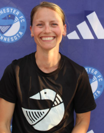 Smiling woman with blonde hair wearing a black sports shirt featuring a white logo of a stylized fish, standing in front of a blue background with a Manchester United crest and Adidas logo.