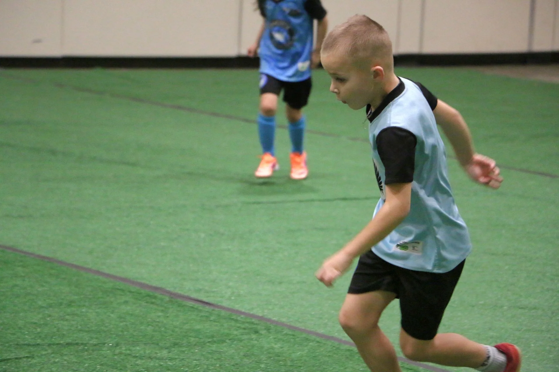 Young boy running and playing indoor soccer on green artificial turf, wearing a light blue jersey with black shorts and red shoes, with a dark-haired child in a blue sports outfit in the background.