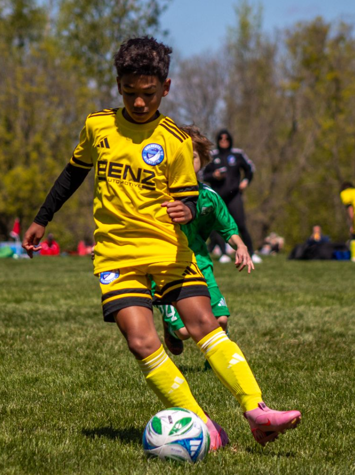 Young boy in yellow soccer uniform kicking a ball during a match on a grassy field.
