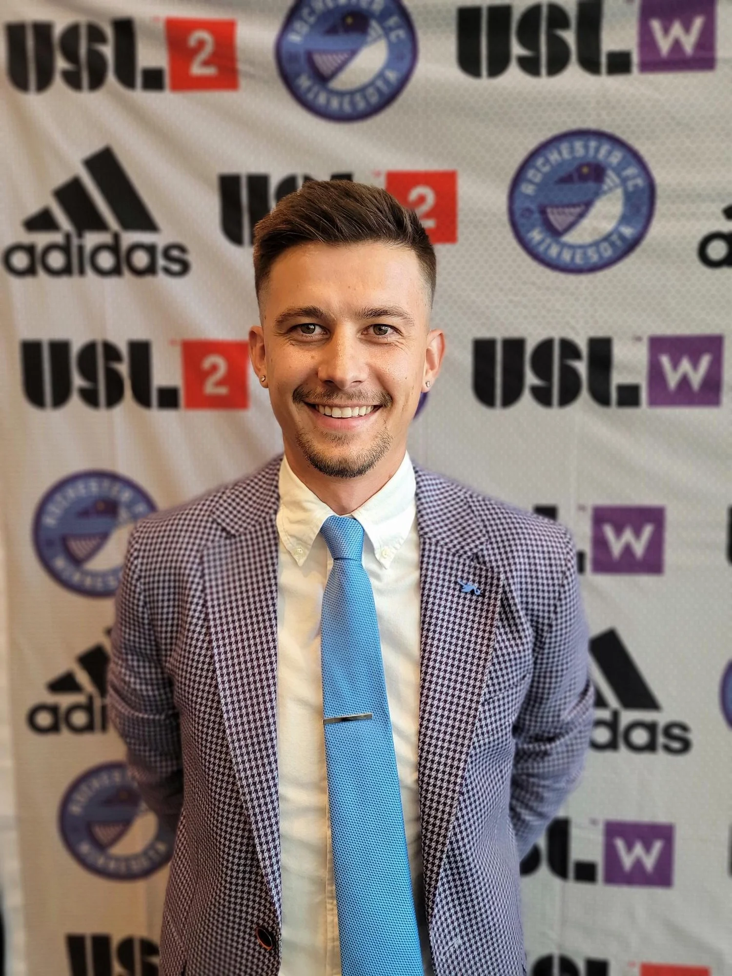 A smiling young man with short dark hair, dressed in a checkered blazer, white shirt, and light blue tie, standing in front of a backdrop with logos including USL 2, Adidas, and Rochester NY.