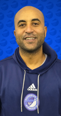 A man with a shaved head and a beard smiling, wearing a dark blue hoodie with the Leicester City football club logo, standing in front of a blue background with a pattern.