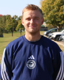 A young man with light brown hair and a beard smiling outdoors. He is wearing a navy blue Adidas sweatshirt with a Seattle Sounders logo. Background includes trees and parked cars.