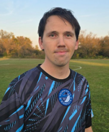 Portrait of a man in a blue and black sports jersey outdoors with trees in the background.