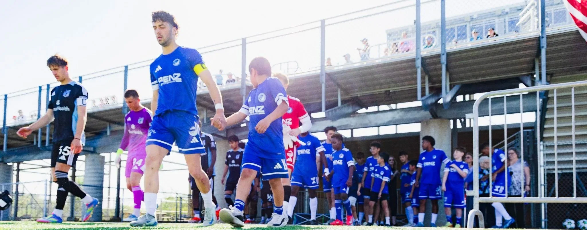 Soccer players walking onto the field before a match, with spectators in the stands and sunny weather.